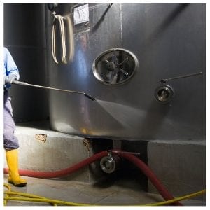 A worker wearing yellow boots and protective clothing uses a high-pressure water hose to clean a large stainless steel industrial tank. Red and yellow hoses are visible on the ground connected to the tank.