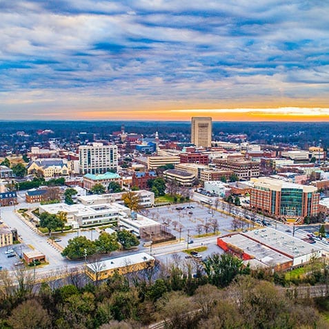Aerial view of an urban cityscape at sunset. The image shows various buildings, including tall structures and smaller establishments, surrounded by trees and roads. The sky is cloudy with hints of sunlight on the horizon.