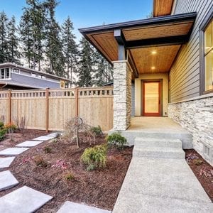 A modern house exterior features a concrete pathway leading to a covered front porch with exposed wooden beams and stone pillars. The yard is landscaped with small plants and a wooden fence encloses the property. Other houses and tall trees are visible in the background.