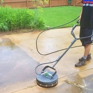A person is using a flat surface cleaner attached to a pressure washer to clean a concrete patio. The left part of the patio is wet and appears clean, contrasting with the right part which is dry and dirty. In the background, a green grassy area and a wooden fence are visible.