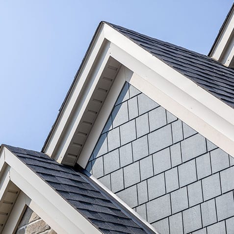 Close-up view of a residential house's roof, featuring three gable roofs with dark grey asphalt shingles and light grey shiplap siding. The angles created by the rooflines are accentuated against a clear blue sky background.