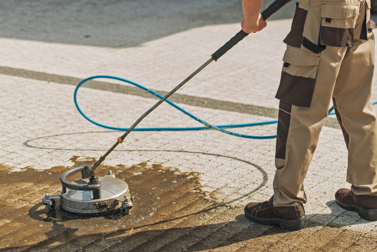 Commercial Concrete Cleaning A worker in beige and brown overalls uses a pressure washer with a circular surface cleaner attachment to clean a paved ground area. A blue hose is connected to the washer, trailing behind the worker. The pavement is partially wet where cleaning has been done.