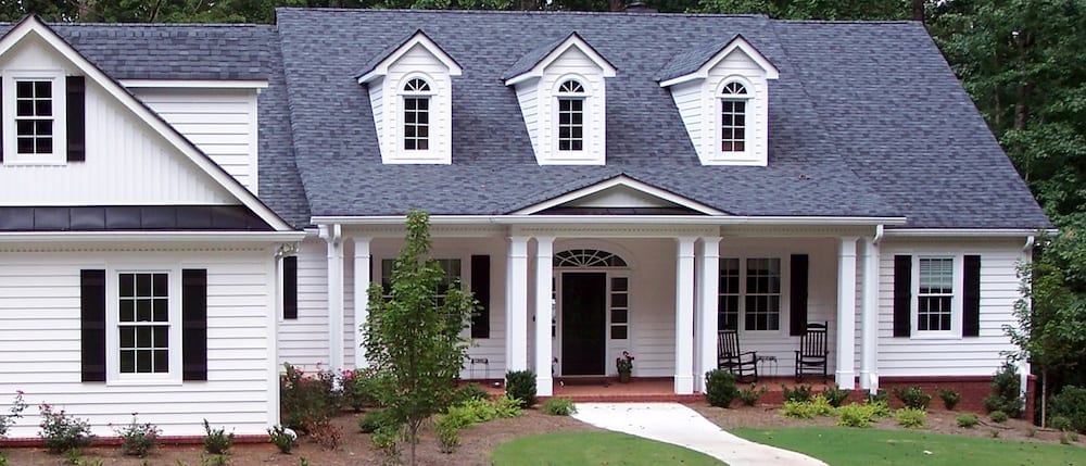 A white, single-story house with blue-gray roofing and three dormer windows on the upper level. It features a covered front porch supported by white columns, a large front lawn with shrubs, and surrounding trees in the background.