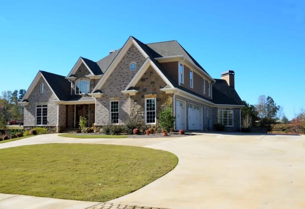 A two-story house with a gray tiled roof and a mix of stone and brick exterior. It features multiple gables, arched windows, a three-car garage, and a well-maintained lawn with shrubs. A large curved driveway leads up to the house. Clear blue sky in the background.