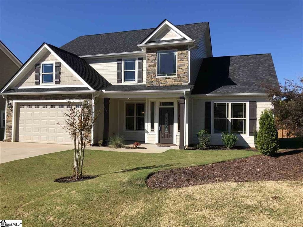 A two-story house with a gray and beige exterior featuring a front porch with columns, stone accents, and two dormer windows. The house has a double garage and a well-maintained front yard with green grass and young shrubbery.