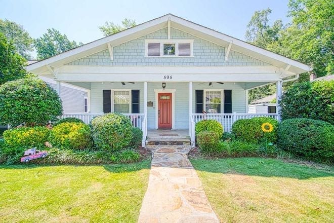A charming single-story house with light blue exterior, white trim, and a red front door. The home has a gabled roof and a spacious front porch with white railings. The yard is neatly landscaped with bushes, flowers, and a small toy car on the left side.