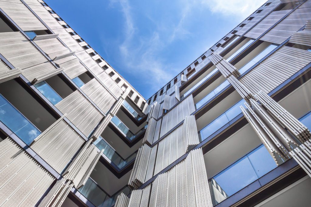 Looking up at a modern, multi-story building with a unique exterior design featuring vertical metallic panels. The balconies have glass railings, and the sky above is clear and blue, enhancing the contemporary architectural style.