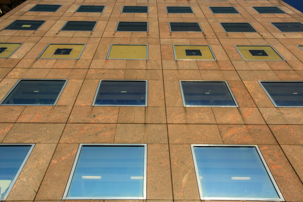 A close-up view of a tall building's facade, showing multiple rows of square windows. The building's surface is made of a reddish-brown stone material, reflecting sunlight and the blue sky above. The windows are arranged in a grid pattern.