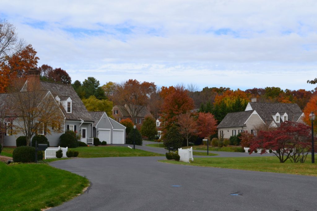 A quiet suburban neighborhood in the fall, with houses featuring gray and white exteriors. The street curves gently, surrounded by green lawns and trees displaying vibrant autumn foliage in shades of red, orange, and yellow. The sky is partly cloudy.